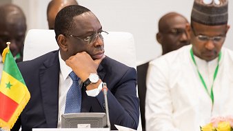 L'image montre un homme assis à une table dans un cadre formel. Il porte un costume sombre et un bracelet montre argenté. Son visage exprime une concentration sérieuse, alors qu'il regarde pensivement devant lui. À sa gauche, il y a un drapeau du Sénégal planté sur la table, indiquant son origine ou son rôle. En arrière-plan, d'autres personnes sont présentes, mais ne sont pas clairement visibles. L'ambiance de la scène suggère un événement officiel, peut-être une réunion ou une conférence de dirigeants.