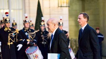 L'image montre deux hommes marchant dans une ambiance officielle. À l'arrière-plan, on peut apercevoir une garde cérémonielle, avec des soldats en uniforme traditionnel, portant des chapeaux distinctifs et tenant des instruments de musique, comme des tambours. Les hommes au premier plan semblent se rendre à une réunion importante, l'un d'eux portant un dossier ou un document. L'éclairage de la scène est doux, créant une atmosphère solennelle et respectueuse.
