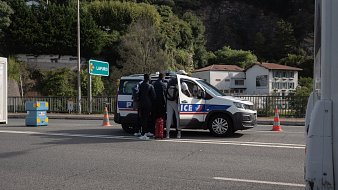 L'image montre une scène sur une route, probablement un contrôle de police. Au centre, on voit une voiture de police de couleur grise avec des marquages bleus et rouges, arrêtée sur la route. Quatre personnes se tiennent près de la voiture, dont deux semblent discuter avec les agents. Ils portent des vêtements sombres et certains semblent avoir des bagages. En arrière-plan, on aperçoit des bâtiments blancs et des arbres qui bordent la route, avec un panneau indiquant "LARRESO". Des cônes de signalisation orange sont disposés autour de la voiture pour sécuriser la zone. L'ambiance paraît calme, bien que la situation soit formelle.
