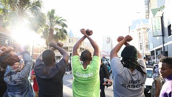 L'image montre un groupe de personnes vues de dos, levant les bras au-dessus de leurs têtes, formant un geste de solidarité ou de protestation. Ils sont sur une rue animée, entourés de bâtiments urbains. On peut apercevoir des palmiers à l'arrière-plan, ce qui ajoute une note de chaleur à l'environnement. La lumière du soleil éclaire la scène, créant une ambiance dynamique. Les personnes portent des t-shirts de différentes couleurs, dont l'un est vert vif. L'ensemble dégage une impression de union et d'engagement collectif.