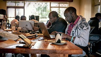 Dans cette image, on voit un espace de travail collaboratif. Plusieurs personnes sont assises autour d'une grande table en bois. Chaque personne utilise un ordinateur portable, principalement des MacBook. Il y a des câbles qui traînent sur la table, ajoutant à l'ambiance de travail intensif. À droite, un homme porte un sweat à capuche gris et écoute des écouteurs, concentré sur son écran. Sur la table, on aperçoit une tasse, peut-être de café, ainsi que quelques feuilles. Les murs sont clairs et de grandes fenêtres laissent entrer la lumière naturelle, créant une atmosphère chaleureuse et productive. En arrière-plan, d'autres personnes sont visibles, toutes absorbées dans leur travail.