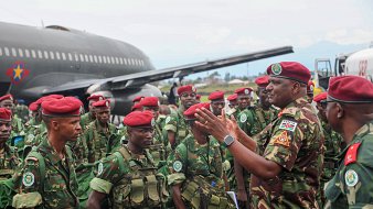 L'image montre un groupe de soldats en uniforme militaire, réunis près d'un gros avion de transport. Les soldats portent des casques et des berets rouges, et ils sont habillés de camouflages verts. L'un des officiers, qui semble donner des instructions ou des encouragements, se tient au centre, gesticulant avec les mains. En arrière-plan, on peut voir l'avion de taille importante, avec des couleurs de camouflage, sur un terrain d'aviation, tandis que le ciel est légèrement nuageux. L'atmosphère est sérieuse, témoignant d'une mobilisation militaire.