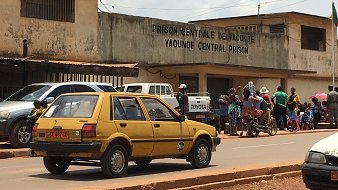 L'image montre l'extérieur de la prison centrale de Yaoundé, au Cameroun. On peut voir un bâtiment sobre avec des murs en béton, portant l'inscription "PRISON CENTRALE DE YAOUNDÉ" en lettres visibles sur la façade. En face de la prison, une route est occupée par une voiture jaune, typique des véhicules urbains. À côté, plusieurs personnes sont rassemblées, certaines discutant, d'autres attendant. Le décor autour est urbain, typique des villes africaines, avec une atmosphère animée. Le ciel est partiellement nuageux, ajoutant une nuance à la lumière de la scène.
