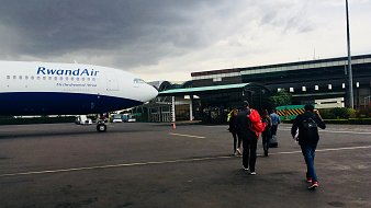 L'image montre un groupe de personnes marchant vers un avion de la compagnie RwandAir, qui est garé sur une aire d'aéroport. L'avion, avec une livrée bleue et blanche, est imposant avec un nez bien visible. Le ciel est nuageux, laissant présager une ambiance légèrement sombre. À l'arrière-plan, on voit un bâtiment d'aéroport avec une architecture moderne. Les voyageurs, portant des sacs à dos et d'autres bagages, avancent vers l'avion, créant un sentiment de départ et d'aventure. Le sol est en béton, et il y a des cônes de signalisation dispersés sur le terrain.