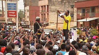 L'image montre une scène de rassemblement dans une rue animée, remplie de nombreuses personnes. Au centre, un orateur, habillé en jaune, se tient sur une plateforme surélevée, tenant un mégaphone et s'adressant à la foule. Les membres de l'audience, qui semblent nombreux et engagés, lèvent souvent leurs téléphones, capturant probablement des moments du discours. En arrière-plan, on aperçoit des bâtiments et une affichage indiquant des prix, ce qui suggère un environnement urbain. L'atmosphère est énergique, marquée par l'enthousiasme et la mobilisation des participants.