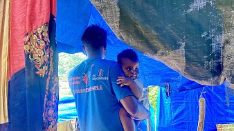 L'image montre un homme portant un enfant dans ses bras. Ils se trouvent sous une tente improvisée faite de tissus aux couleurs vives comme le bleu et le rouge. Le décor semble rural, avec des éléments naturels en arrière-plan. L'homme a un t-shirt bleu avec des inscriptions, et l'enfant, qui regarde légèrement en arrière, semble être de très jeune âge. La scène évoque un moment de tendresse et de protection dans un environnement simple.