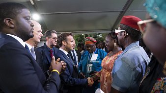 L'image représente un groupe de personnes réunies sous un chapiteau. Au centre, un homme en costume sombre, probablement un leader ou une personnalité importante, est en train de serrer la main d'une femme habillée de façon colorée. On peut apercevoir des visages souriants et une ambiance chaleureuse. D'autres personnes, habillées dans des tenues variées, entourent le couple, regardant avec intérêt. Les arrière-plans sont légèrement flous, mais on ressent une atmosphère de camaraderie et d'échange.