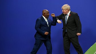 Dans cette image, deux hommes se tiennent sur une scène avec un fond bleu. L'un porte un costume sombre avec une cravate claire, se tenant sur la gauche, et semble sourire en regardant l'autre homme. Ce dernier, sur la droite, est habillé d'un costume noir avec une cravate à motifs et semble également souriant. Les deux hommes portent leurs bras en avant dans un geste de salut amical, touchant légèrement leurs coudes. L'ambiance générale de l'image est dynamique et conviviale, suggérant une interaction positive entre eux.