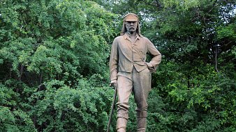 L'image montre une statue en bronze d'un homme qui se tient debout, avec une posture confiante. Il porte des vêtements de style explorateur du XIXe siècle, incluant une chemise à manches longues et un pantalon. Son regard est déterminé et il a une canne à la main, suggérant qu'il pourrait être un aventurier ou un explorateur. La statue est placée sur un socle avec une inscription, et elle est entourée d'une végétation dense, avec des arbres verts en arrière-plan, créant un contraste entre la statue et la nature environnante. L'atmosphère est calme et sereine, reflétant peut-être le respect pour cette figure historique.