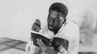 L'image montre un homme assis, plongé dans la lecture d'un livre. Il a la peau foncée et porte une chemise blanche. Dans sa main droite, il tient une cigarette. L'ambiance de la scène est calme, suggérant un moment de réflexion ou d'étude. L'arrière-plan évoque un espace intime, avec des motifs sur le sol qui peuvent donner une impression de confort. L'homme semble absorbé par sa lecture, dégageant une concentration et une sérénité.