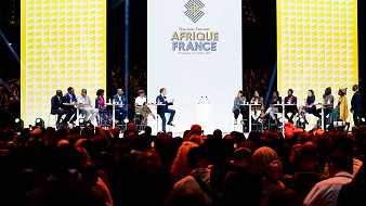 L'image montre une scène d'événement avec plusieurs intervenants assistant à un sommet intitulé "Afrique France". Au centre, un grand écran affiche le logo de l'événement avec une conception graphique moderne. Les intervenants, assis à des tables en cercle, semblent engagés dans une discussion. L'audience est nombreuse et variée, occupant une grande partie de l'espace devant la scène, créant une atmosphère dynamique et interactive. L'éclairage est focalisé sur la scène, mettant en valeur les participants.
