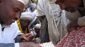 L'image montre un groupe de personnes, principalement des hommes, qui se rassemblent autour d'une table basse sur laquelle se trouve un ancien manuscrit. Un homme au premier plan, portant un habit traditionnel et un turban, montre du doigt une partie du texte. Les autres hommes, qui semblent intéressés, l'observent attentivement. Ils portent également des vêtements traditionnels, et leur expression trahit un mélange de curiosité et de respect. L'ambiance de la scène évoque un moment de partage et de transmission de connaissances, dans un cadre simple mais chargé de culture. En arrière-plan, des éléments naturels et des textures de tissus ajoutent une profondeur à l'atmosphère.