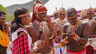 Dans cette image, un groupe de personnes participe à une cérémonie culturelle vibrante. Au premier plan, une femme, vêtue d'une robe traditionnelle colorée avec des motifs, tient un micro et s'adresse à un homme qui se tient à côté d'elle. Cet homme porte un pagne traditionnel et est orné de nombreux accessoires en perles. Ils sont tous deux entourés de plusieurs autres hommes, également en tenue traditionnelle, qui sourient et semblent s'engager dans une conversation joyeuse. Les vêtements sont décorés de motifs colorés et de bijoux en perles, ce qui évoque une atmosphère festive. L'arrière-plan montre des collines verdoyantes, et l'ambiance générale dégage un sens de communauté et de célébration de la culture. L'image respire l'énergie et la convivialité, illustrant des rituels ou des danses traditionnelles.