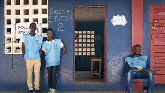L'image montre une scène à l'extérieur d'un bureau de vote. À gauche, deux hommes portent des gilets bleus, probablement identifiant leur fonction d'observateurs ou de travailleurs électoraux. Ils se tiennent debout près de l'entrée, affichant une attitude accueillante. À droite, un autre homme est assis sur une chaise, paraissant détendu. L'environnement est coloré, avec des murs peints en bleu et des fenêtres comportant des grilles. Au-dessus de l'entrée, on peut voir le mot "Polling Station" écrit en anglais, indiquant qu'il s'agit d'un lieu de vote. Le sol est de terre battue, et l'atmosphère semble à la fois tranquille et sérieuse, typique d'un jour d'élection.