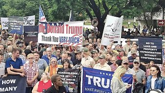 L'image montre une grande foule rassemblée lors d'une manifestation. Les participants tiennent divers panneaux et banderoles. Certains panneaux expriment leur soutien à Donald Trump avec des messages comme "Thank you VERY MUCH" et "MAKE AMERICA GREAT AGAIN". D'autres affichent des revendications ou des slogans liés à des préoccupations politiques. Les manifestants présentent une variété d'âges et de styles vestimentaires, certains portant des chapeaux emblématiques. L'atmosphère semble animée, avec des expressions de passion sur leurs visages. En arrière-plan, des arbres et des panneaux de signalisation sont visibles, créant un cadre urbain pour cette manifestation.