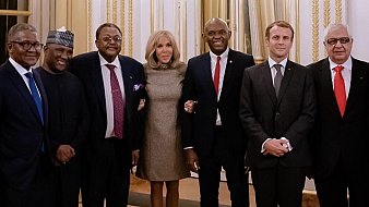 L'image montre un groupe de sept personnes se tenant debout dans une salle élégante, avec des murs clairs et des rideaux drapés. Au centre, une femme souriante porte une robe brillante et se tient entourée de six hommes en costume. Chacun d'eux a un regard chaleureux et engageant. Les hommes varient en teinte de peau et en style vestimentaire, certains portant des cravates, d'autres des vêtements traditionnels. L'ambiance de l'image dégage une atmosphère professionnelle et amicale, suggérant un moment de convivialité ou une rencontre importante.