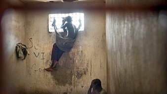 L'image dépeint un environnement sombre et restreint, vraisemblablement une cellule ou une pièce fermée. Au premier plan, deux personnes sont présentes. L'une est assise, visiblement accroupie, la tête entre les mains, adoptant une posture de détresse ou de réflexion. L'autre personne est perchée près d'une fenêtre grillagée, les jambes repliées, enveloppée dans un drap qui semble être une couverture. La lumière qui pénètre par la fenêtre est limitée, créant un contraste entre l'extérieur et l'intérieur sombre de la pièce. Les murs portent des marques, probablement des graffiti, ce qui suggère un environnement négligé. L'atmosphère générale est lourde et empreinte de mélancolie, reflétant un sentiment d'isolement et de désespoir.