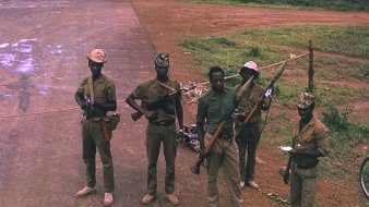 L'image montre un groupe de cinq soldats en uniforme, debout sur une route non pavée. Ils portent des chapeaux, et chacun d'eux tient une arme à feu. Le sol est poussiéreux, avec quelques zones humides visibles. En arrière-plan, on voit une végétation luxuriante, typique d'un environnement tropical. Les soldats affichent des postures sérieuses, et certains d'entre eux paraissent concentrés sur leur environnement. Cette scène suggère un contexte de sécurité ou de patrouille.