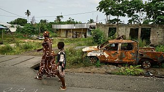 L'image montre une scène d'une rue dans un environnement rural ou semi-urbain. Sur le trottoir, une femme vêtue d'une longue robe ornée de motifs blancs marche aux côtés d'un jeune garçon. Ils avancent côte à côte, probablement en direction d'un endroit précis. En arrière-plan, on peut voir un paysage verdoyant avec des arbres, ainsi que des bâtiments partiellement construits, évoquant une ambiance de quartier en développement. À droite, un vieux véhicule, rouillé et en piteux état, est abandonné sur le côté de la route, entouré de végétation. Le ciel est nuageux, suggérant qu'il pourrait faire chaud et humide. L'ensemble dégage une atmosphère à la fois paisible et un peu nostalgique.