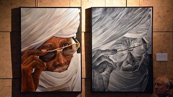 L'image présente deux tableaux accrochés sur un mur, représentant le même homme, mais dans des styles différents. À gauche, le portrait est en couleurs vives : un vieil homme porte un turban blanc et des lunettes de soleil. Son regard est pensif, avec une expression de sagesse. À droite, l'œuvre est en noir et blanc, offrant un contraste fort. Les détails des rides et des textures de la peau sont accentués, donnant une sensation de profondeur. En bas à droite de l'image, on aperçoit une silhouette d'un visiteur qui observe les œuvres. Le fond du mur est constitué de panneaux en bois clair, ajoutant une dimension chaleureuse à l'espace d'exposition.