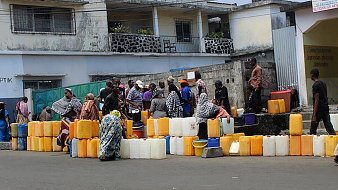 L'image montre une scène animée dans une rue. Des groupes de personnes se rassemblent autour de plusieurs conteneurs en plastique de différentes tailles, souvent de couleur orange ou jaune. Ces conteneurs sont alignés sur le sol. Les personnes, principalement habillées de vêtements traditionnels, semblent attendre leur tour pour remplir les récipients d'eau. En arrière-plan, on distingue des bâtiments aux façades simples, certains avec des balcons. L'ambiance est celle d'une activité quotidienne, soulignant l'importance de l'eau dans la vie des gens.