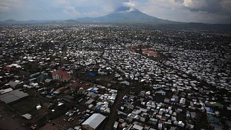 Imaginez une vaste étendue urbaine vue d'en haut, où des milliers de toits en tôle ondulée et en matériaux divers se mêlent pour créer un paysage dense. Les bâtiments semblent serrés les uns contre les autres, formant un réseau labyrinthique. Au loin, un majestueux volcan émerge à l'horizon, avec des sommets nuageux qui soulignent sa présence imposante. Le ciel présente des teintes variées de gris et de bleu, projetant une lumière diffuse sur la scène. En bas, des routes sont visibles, animées de véhicules et de personnes, ajoutant une sensation de vie à cet environnement. Cette image évoque à la fois la densité urbaine et la force tranquille de la nature en toile de fond.