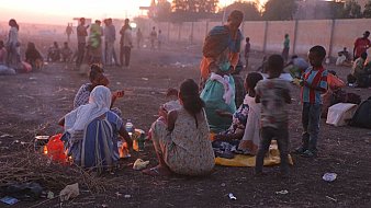 L'image montre une scène de vie en plein air, probablement à la tombée de la nuit. Au premier plan, plusieurs femmes et enfants sont assis sur le sol, entourés de quelques objets personnels et de restes de nourriture. Les femmes, habillées de vêtements colorés, semblent préparer un repas autour d'un feu. En arrière-plan, la lumière du crépuscule illumine légèrement la scène, et de nombreuses personnes peuvent être aperçues se déplaçant dans un environnement poussiéreux. L'atmosphère dégage à la fois une certaine convivialité et une sensation de précarité.