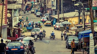 L'image dépeint une rue animée d'une ville. On y voit des bâtiments à plusieurs étages, certains colorés, avec des toits en tôle. La rue est bordée de voitures et de deux roues, et on aperçoit des personnes se déplaçant sur le trottoir. Certaines personnes marchent, d'autres se déplacent à moto. Des parasols colorés sont installés sur le trottoir, offrant de l'ombre, et il y a des fils électriques qui traversent l'espace. L'atmosphère est vivante et dynamique, reflétant le quotidien d'une ville.