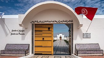 Imaginez un lieu paisible et accueillant, avec une grande porte jaune ornée de motifs géométriques. Cette porte s'ouvre sur un jardin magnifique, symbolisant la beauté et la diversité de la nature. Deux bancs placés de chaque côté de l'entrée, recouverts de carrelage coloré, invitent à la détente. Au-dessus de la porte, une inscription en arabe et en français nous invite à découvrir ce jardin, intitulé "Jardin d'Afrique, Jardin du Paradis". De chaque côté, un drapeau flotte, ajoutant une touche de couleur vive à la scène. Le ciel est lumineux et parsemé de nuages, apportant une lumière douce qui souligne la sérénité de cet endroit. L'ambiance générale est calme et harmonieuse, évoquant un espace de ressourcement et de contemplation.