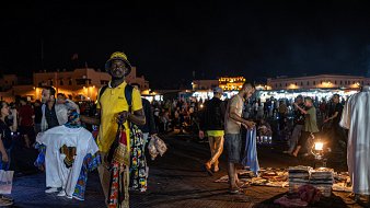Dans cette image prise la nuit dans un marché animé, on voit un homme au premier plan portant un t-shirt jaune et un chapeau. Il tient plusieurs vêtements, avec des motifs colorés, dans chaque main. L'arrière-plan est rempli de personnes se déplaçant dans le marché, créant une ambiance vivante et dynamique. On peut également apercevoir des lumières brillantes provenant de stands qui éclairent la scène, ajoutant à l'animation nocturne du lieu. La texture du sol semble être en bois ou en pavé, créant un environnement chaleureux. L'air est rempli des sons de conversations et de l'agitation du marché.