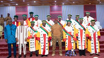 L'image montre un groupe de personnes posant ensemble dans un cadre formel. Au centre, il y a un homme en tenue militaire, entouré par un groupe de personnes habillées en robes hautes avec des motifs distinctifs et des chapeaux colorés. Les vêtements semblent refléter une certaine tradition ou un statut élevé, avec des éléments de couleur rouge, vert et jaune. Les membres du groupe arborent aussi des écharpes qui traversent leur poitrine, ajoutant une touche de distinction. En arrière-plan, il y a des décorations murales et une scène de cérémonie qui donne une atmosphère solennelle à l'ensemble. Le sol est recouvert d'un tapis rouge, contribuant à l'élégance du moment.