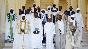 L'image montre un groupe de personnes habillées en vêtements traditionnels, rassemblées dans un espace élégant. Ils sont principalement vêtus de robes blanches, avec des éléments colorés comme des capes vertes et bleues. Les hommes portent des turbans et des chapeaux, ajoutant à la richesse de leurs tenues. L'arrière-plan est un intérieur lumineux et spacieux, avec des colonnes et des décorations architecturales. Le groupe semble coordonné et solennel, probablement pour une occasion spéciale.