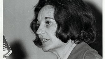 L'image montre une femme avec des cheveux foncés, mi-longs et légèrement ondulés. Elle est en profil, parlant près d'un microphone. Son expression est concentrée, et on peut deviner une certaine passion dans son discours. Elle porte une veste simple, et l'arrière-plan est flou, ce qui met l'accent sur son visage et son expression. L'éclairage est doux, soulignant les traits de son visage.