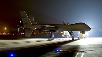 L'image montre un drone de surveillance, typiquement de type militaire, stationné sur une piste d'atterrissage. La scène est nocturne, éclairée principalement par des lumières artificielles qui révèlent les contours du drone. Le drone a une forme allongée et est équipé d'une hélice à l'arrière. Il repose sur un châssis avec des supports, et il y a des lumières au sol qui peuvent délimiter la zone ou indiquer l'emplacement. L'ambiance est sérieuse et technologique, évoquant des missions de surveillance ou de reconnaissance.