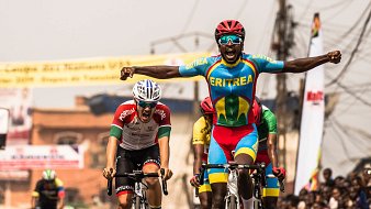 L'image montre un moment dynamique d'une course de cyclisme. Au premier plan, un coureur, vêtu d'un maillot multicolore représentant l'Érythrée, lève les bras en signe de victoire, affichant une expression de joie et d'excitation. En arrière-plan, un autre cycliste, portant un maillot aux couleurs du Portugal, semble montrer des signes d'effort intensif, presque à la limite de l'épuisement. On aperçoit également d'autres participants et une foule enjouée de spectateurs, créant une ambiance festive autour de cet événement sportif. Le décor est vibrant, évoquant l'énergie et la compétitivité de la course.
