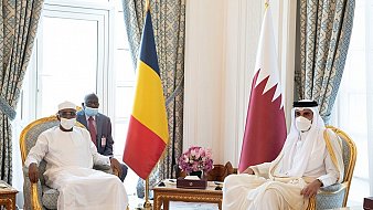 L'image montre une réunion formelle entre deux dirigeants. À gauche, un homme porte une tenue traditionnelle blanche et un chapeau, symbole de son pays. À sa droite, un autre homme est habillé d'une robe longue beige et d'un couvre-chef traditionnel. Entre eux, une table basse en bois contient des objets décoratifs. En arrière-plan, on peut voir des drapeaux : celui du Tchad à gauche et celui du Qatar à droite. La décoration des murs et des rideaux est élégante, avec des motifs riches et des couleurs apaisantes. L'atmosphère est solennelle et professionnelle.
