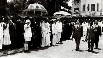 L'image montre un groupe de personnes rassemblées en extérieur, probablement lors d'un événement officiel. Au centre, des hommes en costumes formels marchent, entourés de dignitaires et de membres du public. Certains portent des uniformes militaires, tandis que d'autres sont habillés de manière civile. Il y a également des parapluies décoratifs en arrière-plan. L'atmosphère semble solennelle, et l'environnement suggère une atmosphère tropicale avec des arbres en arrière-plan. La scène donne une impression de cérémonie et de célébration.