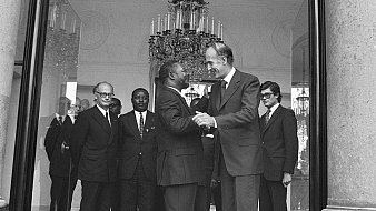 L'image montre un groupe d'hommes en costume, réunis dans un grand hall bien éclairé. Au centre, deux hommes se serrent la main, affichant des expressions de cordialité. L'un est légèrement plus âgé, avec des cheveux grisonnants, tandis que l'autre est plus jeune. En arrière-plan, plusieurs autres hommes les observent, tous vêtus de costumes sombres, ce qui accentue l'atmosphère formelle de la rencontre. Le décor est élégant, avec un grand lustre suspendu au plafond et des colonnes en pierre qui ajoutent une touche de sophistication.