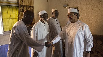 L'image montre un groupe de quatre hommes dans une pièce intérieure. Ils portent des vêtements traditionnels : des turbans et des vêtements clairs, souvent en coton. Un des hommes, sur la droite, est en train de serrer la main d'un autre homme, tandis que deux autres les regardent avec un air amical. La lumière entre par une fenêtre derrière eux, créant une atmosphère chaleureuse. Les murs sont texturés, avec un éclairage doux, et l'ensemble donne une impression de convivialité et de respect mutuel.