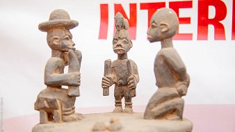 L'image présente une sculpture en bois représentant trois figures humaines stylisées. Au centre, une figure masculine, aux traits marqués, tient un objet dans les mains, probablement un outil ou un symbole culturel. Il est entouré de deux autres sculptures, l'une à gauche, qui porte un chapeau large, et l'autre à droite, avec une coiffure distinctive. Les personnages semblent engagés dans une conversation ou un échange, ce qui suggère une interaction sociale. Les détails des sculptures, comme les expressions des visages et les postures, témoignent d'un art tribal riche en histoire et en culture. Le fond de l'image est flou, mettant l'accent sur les sculptures elles-mêmes.