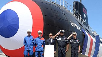 L'image montre un sous-marin de couleur noire avec un grand cercle rouge et bleu sur son côté. Devant le sous-marin, il y a quatre personnes en tenue de travail, dont deux en bleu et deux en uniforme noir. Elles se tiennent debout, affichant des expressions sérieuses. En arrière-plan, le ciel est clair et bleu, donnant une ambiance ensoleillée à la scène. Au centre, une plate-forme sur laquelle un homme en costume parle, probablement lors d'une cérémonie liée au sous-marin. Cela évoque une atmosphère de célébration ou d'inauguration d'un nouveau bateau.