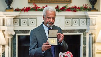 L'image montre un homme âgé, avec des cheveux poivre et sel et une barbe, qui porte un costume gris. Il se tient fièrement devant une cheminée décorée, tenant une médaille en or dans un écrin, qu'il montre à la caméra. En arrière-plan, on peut distinguer une décoration florale avec des fleurs disposées sur une table, créant une ambiance chaleureuse et élégante. La pièce est bien éclairée et a un air prestigieux, suggérant une occasion spéciale.