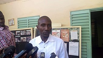 Dans cette image, un homme se tient devant un mur avec des panneaux d'affichage. Il porte une chemise blanche et semble parler à plusieurs journalistes qui l'entourent, tenant des microphones et des appareils d'enregistrement. Les panneaux d'affichage derrière lui contiennent des informations, des photos et des affiches. L'ambiance paraît être celle d'une conférence de presse ou d'un événement public où des informations sont partagées.