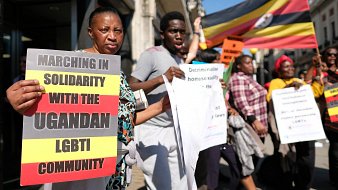 Dans cette image, un groupe de personnes participe à une marche de solidarité en faveur de la communauté LGBTI ougandaise. Les manifestants tiennent des pancartes avec des messages tels que "Marchons en solidarité avec la communauté LGBTI ougandaise" et "Décriminalisez l'homosexualité". L'ambiance est énergique et déterminée, et le groupe affiche une grande diversité. En arrière-plan, on peut voir un drapeau de l'Ouganda flapper, créant un contraste visuel avec les affiches et les vêtements des participants. Les personnes expriment un fort engagement pour les droits humains et l'égalité.