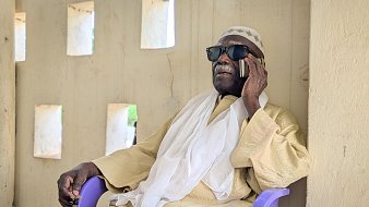 L'image montre un homme assis sur une chaise en plastique violette, contre un mur de couleur beige avec des fenêtres rectangulaires qui laissent passer une lumière douce. Il porte des lunettes de soleil noires et un vêtement traditionnel de couleur claire, accompagné d'un châle blanc drapé sur ses épaules. L'homme est en train de parler au téléphone, son visage exprime la concentration et la sérénité. L'environnement est calme, évoquant un moment de détente dans un cadre paisible.