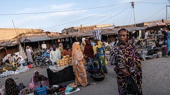 Dans cette scène animée d'un marché, on peut ressentir une atmosphère vibrante. Le sol est recouvert de sable, et plusieurs stands sont disposés tout autour. Des gens sont rassemblés, certains vêtus de vêtements colorés, offrant divers produits alimentaires, tels que des fruits et des pâtisseries. Les femmes, en particulier, attirent l'attention avec leurs robes chatoyantes et leurs voiles. On peut aussi entendre des murmures de conversations, des rires et le bruit de petits véhicules, comme des motos, qui se déplacent à travers le marché. L'air est imprégné d'odeurs variées de nourriture épicée et de douceur, créant une expérience sensorielle unique.