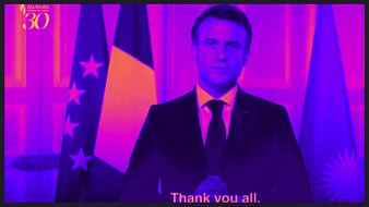 L'image montre un homme en costume sombre, debout devant un arrière-plan aux tons violets et roses. Il a une expression sérieuse et est placé entre deux drapeaux : un drapeau tricolore français à gauche et un autre qui pourrait représenter l'Argentine à droite. Ses mains sont jointes devant lui, comme s'il allait s'exprimer. En bas de l'image, on peut lire un texte en anglais qui dit "Thank you all", indiquant qu'il s'adresse à un auditoire. L'ensemble de la scène dégage une atmosphère formelle et solennelle.