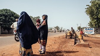 Dans cette image, nous sommes sur une route dans un environnement urbain. Sur le côté gauche, deux femmes portant des vêtements traditionnels de couleur sombre se tiennent ensemble. L'une d'elles a un sourire et semble discuter. Elles sont entourées de terre brune, et l'atmosphère est calme. Sur le côté droit, un homme est assis près d'un petit chariot, tandis qu'un panneau indique « GAGI », signalant un commerce local. On peut apercevoir d'autres personnes se déplaçant à pied ou à bicyclette sur la route, soulignant la vie active de ce lieu. Au fond, des arbres verdoyants ajoutent une touche de nature à l'environnement urbain.