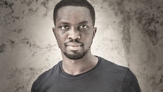 Cette image présente un homme debout, portant un t-shirt noir. Il a la peau foncée et un style de coiffure court. Son expression est sérieuse, et il regarde directement vers l'objectif avec une intensité qui attire l'attention. Le fond de l'image est texturé et neutre, mettant en valeur son visage et son corps. La lumière éclaircit certaines zones, ajoutant de la profondeur et du contraste à l'ensemble.