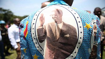 Cette image montre une personne portant un grand vêtement aux motifs colorés et vifs. Le dos du vêtement est orné d'une grande impression d'une personne en costume, levant la main dans un geste de salut. Autour de l'image, on peut voir des étoiles et des motifs géométriques. La date "1961-01-17" est inscrite, indiquant probablement une signification importante liée à l'image. En arrière-plan, on aperçoit un groupe de personnes, suggérant un événement ou une réunion en extérieur sous un ciel bleu.